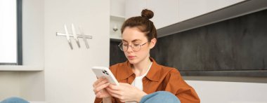 Gözlüklü esmer kadının portresi, evde cep telefonuyla oturuyor, akıllı telefonunu kullanıyor ve gülümsüyor, mesaj okuyor..