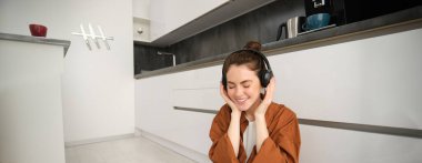 Evdeki mutfak zemininde güzel bir kadın modelin portresi, kablosuz kulaklıkla müzik dinliyor, gülümsüyor ve mutlu görünüyor..