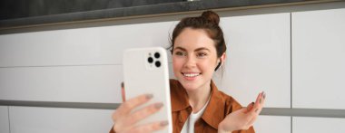 Güzel gülümseyen bir kadının portresi, akıllı telefondan arkadaşınla görüntülü konuşma, mobil uygulama üzerinden çevrimiçi sohbete bağlanma, mutfak zemininde oturma.
