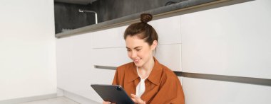 Evde çalışan ya da ders çalışan genç bir kadının portresi dijital tablet kursunu izliyor, mutfak zemininde oturuyor, gülümsüyor ve mutlu görünüyor. İnsanlar ve yaşam tarzı kavramı