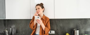 İnternetten akıllı telefon uygulaması kullanan, düşünen yüzüyle mutfakta oturan, elinde cep telefonu tutan bir kadının portresi..