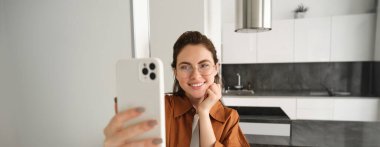 Gözlüklü güzel bir kadın evinde selfie çekiyor, akıllı telefon kullanıyor, fotoğraf için poz veriyor, cep telefonuyla duruyor ve gülümsüyor..