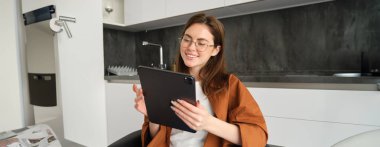 Gözlüklü genç bir kadının portresi, dijital tablete bakıyor, evden çalışıyor, uzak sınıflara bağlanıyor veya internetten video seyrediyor..
