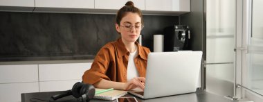 İş ve yaşam dengesi kavramı. Gözlüklü genç bir kadın, evden çalışıyor, dizüstü bilgisayarda yazı yazıyor, bilgisayar ödevi yapıyor, mutfakta oturuyor, serbest çalışıyor..