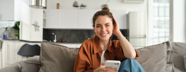 Güzel, gülümseyen bir kadın model akıllı telefonuyla koltukta oturuyor, kameraya mutlu bakıyor, sosyal medyaya kayıyor, mesajlaşıyor, online alışveriş yapıyor..