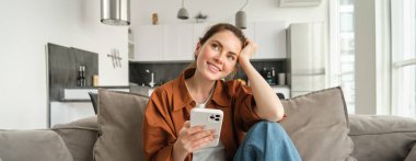 Gülümseyen esmer kadın akıllı telefonuyla koltukta oturuyor, düşünceli görünüyor, cep telefonu uygulamasını kullanırken düşünüyor, başvuru siparişi veriyor, online alışveriş yapıyor ve evde dinleniyor..