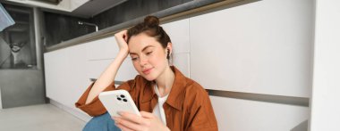 Gülümseyen güzel bir kadının portresi mutfak zemininde oturuyor, cep telefonuyla sosyal medyayı kaydırıyor, kablosuz kulaklıkla müzik dinliyor..