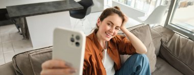Güzel mutlu kızın portresi kanepede selfie çekiyor. Kanepesinde poz veren, sosyal medya için fotoğraf çeken bir kadın akıllı telefonuyla elini uzatıyor..