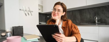 Şirin genç bir kadının portresi, serbest çalışan bir öğretmen, mutfakta gözlük takıyor, dijital tablet tutuyor, insanlara online öğretiyor, derslere bağlanmak için alet kullanıyor, uzaktan çalışıyor..