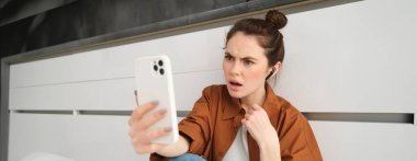 Şok olmuş, hayal kırıklığına uğramış bir kadın portresi, akıllı telefon ekranına bakıyor, telefondan kötü haberler duyuyor, kablosuz kulaklıkla konuşuyor..