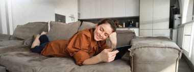 Güzel genç bir kadının portresi, evinde kanepede dinleniyor, dijital tabletle kanepede uzanıyor, kitap okuyor ya da aletin üzerinde video izliyor..