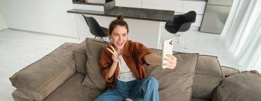 Şirin gülümseyen kadın modelin portresi, akıllı telefon uygulamasına selfie çekiyor, sosyal medyaya fotoğraflar yüklüyor, cep telefonu kamerası kullanıyor, evdeki kanepede poz veriyor..