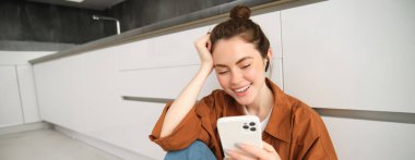 Kablosuz kulaklıklı genç bir kadının portresi, mutfak zemininde oturuyor, akıllı telefon kullanıyor, favori şarkılarla çalma listesi yapıyor, müzik dinliyor..