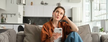 Akıllı telefonlu kanepede oturan sevimli, meraklı bir kadının portresi kafasını kaldırıp düşünüyor, cep telefonu uygulamasından internetten alışveriş yapıyor..