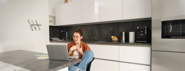 Evden çalışmak, uzaktan iş kavramı. Genç bir kadın mutfakta oturuyor, dizüstü bilgisayar kullanıyor, görüntülü sohbete katılıyor, çevrimiçi dersler veriyor, bilgisayarda müşteriyle konuşuyor..