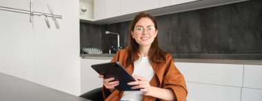 E-öğrenme ve uzak iş yeri konsepti. Gözlüklü genç bir kadın, mutfakta okuyan bir öğrenci, dijital tablet okuyor, projesine bakıyor, online toplantıya bağlanıyor..