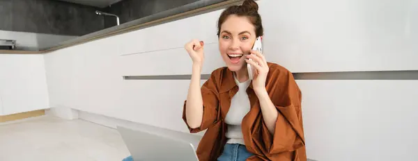Happy girl sits on floor, receives great news over the phone and triumphs, celebrates and feels excited hearing positive information.