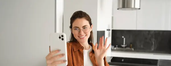 Portrait of stylish beautiful woman at home, video chats on smartphone, connects to online meeting from her living room, lifestyle social media blogger records a video on mobile phone.
