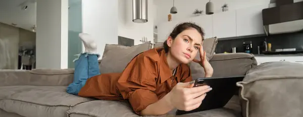 Portrait of woman lying on couch at home, holding digital tablet and looking thoughtful, frowning while thinking and looking aside, making decision, purchase online on gadget.