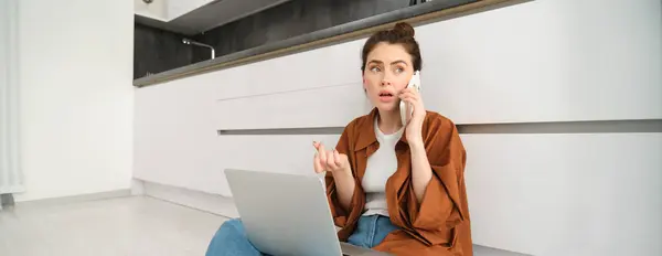 Woman with concerned, worried face, making a phone call, sitting on floor with laptop and having difficult conversation over telephone.