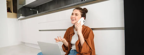 Cheerful smiling woman sits on floor in the kitchen with laptop, makes a phone call, talks to friend and laughs during conversation.