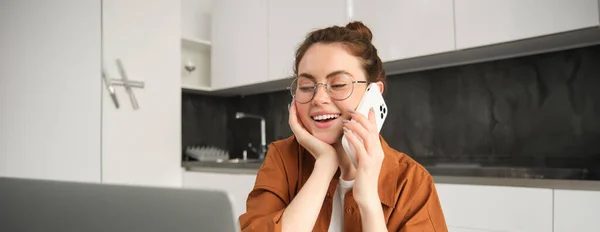 Portrait of beautiful young woman managing her own business from home, freelancer making phone call, sitting in kitchen with laptop and smiling.