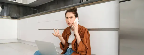 Woman with frustrated and confused face talking on mobile phone, shrugging and frowning, hear bad news over the telephone, sits on floor with laptop, calls customer support.