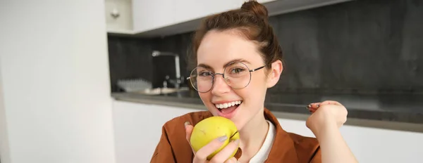 Close-up portrait of brunette woman at home, wearing glasses, eating apple in the kitchen and smiling, biting fruit.