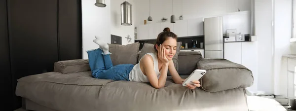 stock image Portrait of smiling, happy young woman resting at home, resting on couch with smartphone, using mobile phone application.