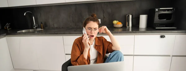 Portrait of woman with shocked face, receives concerning bad news over the phone conversation, sitting at home with laptop, working, talking to someone on telephone.