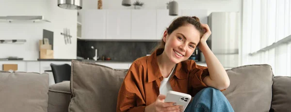 Portrait of cute young smiling woman, sitting on sofa with mobile phone, relaxing at home and using smartphone, scrolling social media, buying online, ordering something on application.
