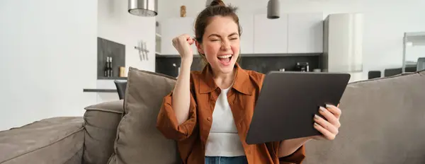 Happy, enthusiastic young woman winning on video game, looking at digital tablet and celebrating victory, triumphing and laughing, sitting on couch at home.