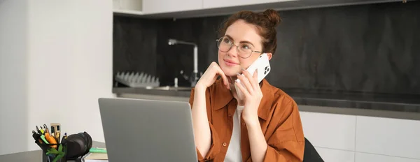 Portrait of young self-employed woman, entrepreneur working from home, freelancer calling client. Girl making an order, talking to someone on phone, sitting in kitchen with laptop.