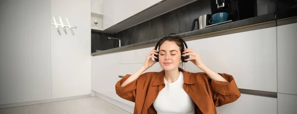 Cute brunette woman on kitchen floor, wears headphones, listens music and smiles, enjoys sound quality.