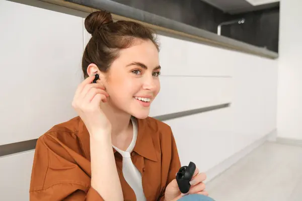 Image of modern brunette girl, sits on kitchen floor, listens to music in wireless headphones, holding earphones and case in hands, looking aside.