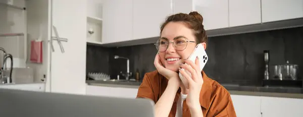 Portrait of beautiful young woman managing her own business from home, freelancer making phone call, sitting in kitchen with laptop and smiling.