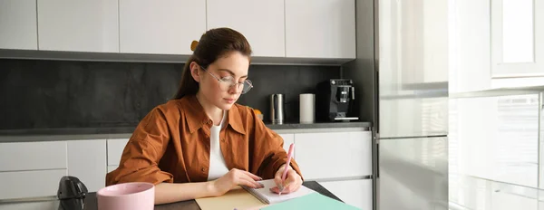 stock image Portrait of woman looking concentrated, writing down notes, doing homework in kitchen, drinking coffee, studying at home.