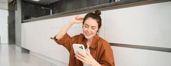 Portrait of happy young beautiful woman, listening music in wireless earphones, using smartphone, watching videos on mobile phone with headphones on.