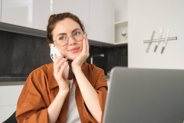 Evden çalışan güzel gülümseyen bir kadının portresi, cep telefonuyla konuşan, müşteriyi arayan, serbest çalışan bir iş kadını, dizüstü bilgisayar ve akıllı telefon kullanarak mutfakta iş yeri kuruyor..