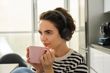 Gülümseyen esmer kadının yakın portresi, öğrenci çayını içiyor ve kulaklıkla müzik veya kitap dinliyor, mutfakta oturuyor ve dinleniyor..
