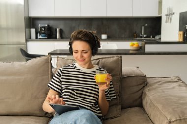 Modern kızın portresi, portakal suyu içmek, dijital tablette videolar izlemek, oturma odasındaki kanepede oturmak..