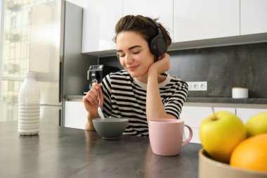 Güzel genç bir bayan model, mutfakta kahvaltı yapıyor, mısır gevreğini sütlü yiyor, gülümsüyor ve kahve içiyor, müzik dinliyor ya da kablosuz kulaklıkla podcast yapıyor..