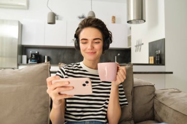 Genç bir kadın akıllı telefon uygulamasında videolar izliyor, kulaklıkla çay içiyor, kanepede oturuyor, evinde rahat vakit geçiriyor..