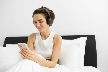 Yatağa giden genç bir kadının portresi, kulaklık takan, akıllı telefon kullanan, podcast ya da istiridye dinleyen, kulaklıkla müzik dinleyen..