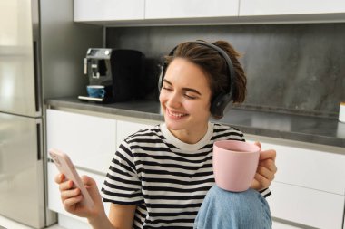 Çay içen ve kulaklıkla müzik dinleyen genç bir kadının portresi, mutfakta yemek yerken sosyal medyayı kaydırmak, mutlu bir şekilde gülümsemek..