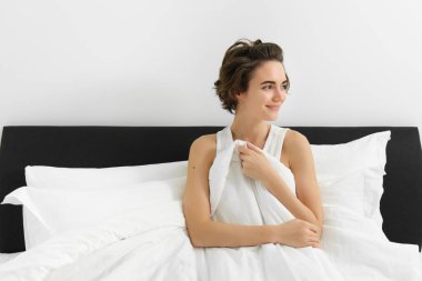 Dağınık saçlı genç esmer kadın, yatağında uyanıyor, şehvetli, uykulu bir gülümsemeyle yan yana oturuyor, keten beyazı çarşaflarla kendini örtüyor..