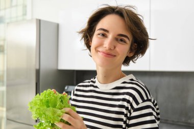 Gülümseyen esmer kadının yakın portresi, mutfakta duruyor, yeşil marul yaprağı tutuyor, kameraya mutlu bakıyor, diyette sağlıklı yiyecekler yiyor, salata yapıyor..