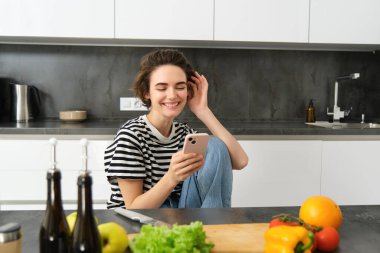 Gülümseyen, gülen ve akıllı telefon kullanan genç bir kadının portresi yemek pişirirken, akıllı telefon uygulamasında yemek tarifleri ararken, evdeki mutfağında sebzelerden yemek hazırlarken..