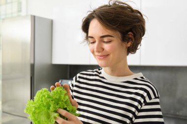 Mutfakta elinde taze yeşil marul yaprakları, salata için sebze hazırlayan sağlıklı vejetaryen yemeği pişiren genç bir kadının portresi..