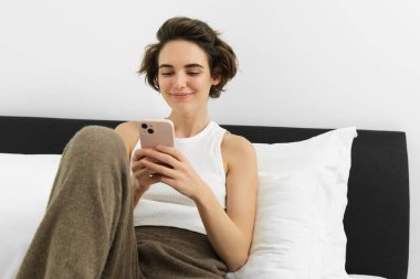 Genç güzel bir kadının portresi yatakta uzanıyor, yatak odasında dinleniyor, mesajlaşıyor, cep telefonu kullanıyor, akıllı telefonu tutuyor ve gülümsüyor, sosyal medyada kayıyor, evde rahat zaman geçiriyor..